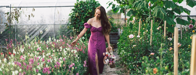 Woman walking through a lush flower garden, gently touching blooming plants and holding a basket of fresh flowers in a natural outdoor setting
